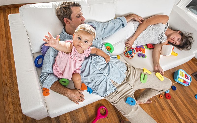 Auf dem Bild liegt eine Frau zwischen Kinderspielzeug auf dem Sofa und schläft - ein Mann hat ein Kleinkind im Arm das putzmunter ist und er liegt schräg auf der Coach ung ist auch am schlafen oder zumindest hat er die Augen zu. Die Coach ist weiß das Kind ist etwa 1-1 1/2 Jahre alt mit einem rosa Kleid. Der Vater hat ein blaues Hemd und eine beige Hose an. Elke Maria Becker Paartherapie - Paarberatung Niedernberg, Aschaffenburg, Miltenberg, Walldürn