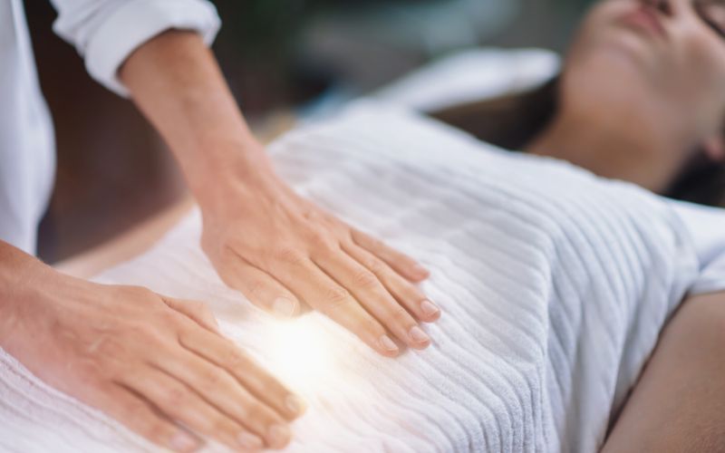 Reiki Anwendung, Frau liegt auf dem Rücken und Reiki Praktizierende hat beide Hände auf den Bauch gelegt und gibt Reiki Energie, Bauch leuchtet im Licht, Elke Maria Becker, Niedernberg bei Aschaffenburg und online