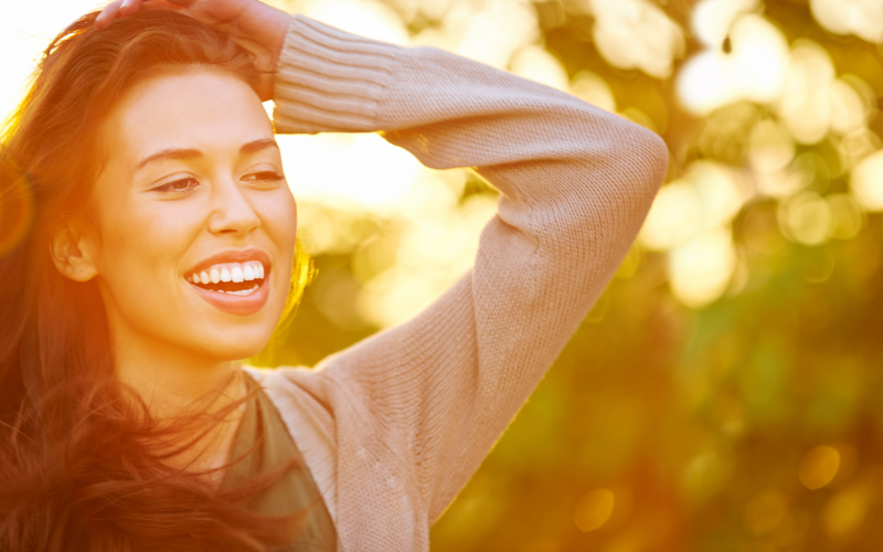 Selbstliebe, Frau mit langen braunen Haaren strahlt und hat die linke Hand am Hinterkopf. im Hintergrund sind Bäume und Sonnenlicht, Reiki in Niedernberg bei Aschaffenburg, Elke Maria Becker, Reiki Meisterin und Lehrerin