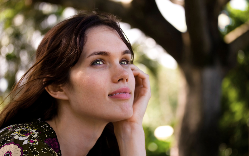 Frau mit langen braunen Haaren hat Kopf auf linke Hand gestützt und schaut überlegend. Im Hintergrund ist ein Baum, Reiki, Elke Maria Becker, Niedernberg bei Aschaffenburg 